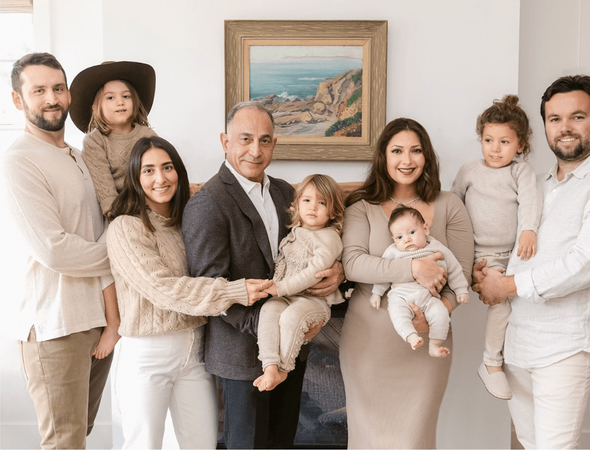 Mo Honarkar with his daughters, their husbands and his grandchildren, gathered in a light‑filled room beneath a coastal landscape painting.